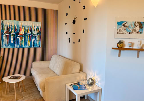 Soggiorno con divano beige, tavolino bianco, orologio da parete nero, pianta in vaso, e due quadri appesi.