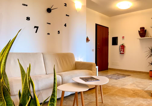 Soggiorno con divano beige, tavolini bianchi, pianta verde, orologio a muro moderno e porta d'ingresso.