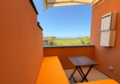 Balcone con pareti arancioni, tavolo di legno e panca arancione, vista su alberi e cielo azzurro.