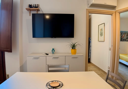 Sala con tavolo bianco, televisore a schermo piatto su una parete, mobile con decorazioni, porta aperta che conduce a una camera da letto con letto giallo.