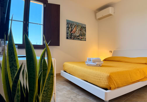 Camera da letto con letto matrimoniale coperto da un copriletto giallo, finestra aperta, pianta verde in primo piano e quadro appeso alla parete.