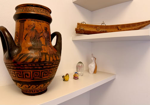 Un vaso decorato su uno scaffale bianco, accanto a piccoli oggetti e una scultura a forma di barca su un altro scaffale.